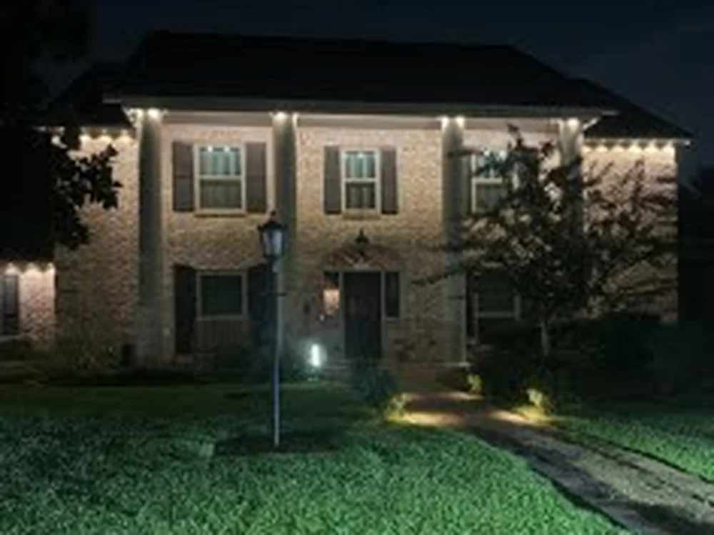 Real BrightSky install — two-story brick home with warm-white permanent lighting outlining the eaves, entry, and a softly lit lawn at night